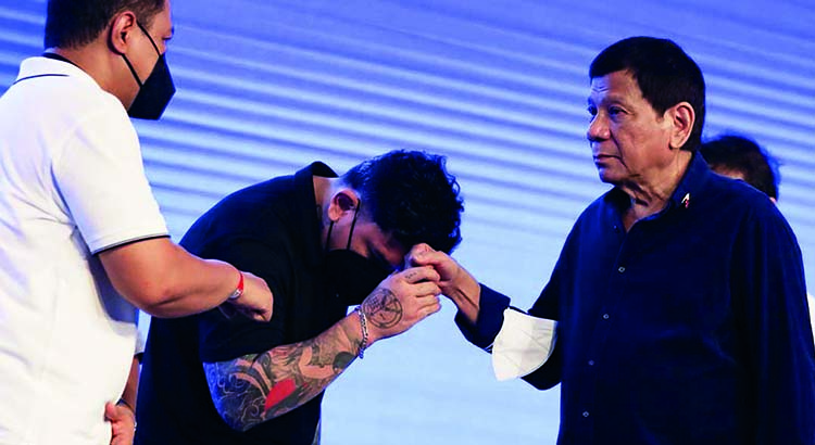 Sebastian „Baste“ Duterte verneigt sich und führt die Hand seines Vaters, Präsident Rodrigo Duterte, an seine Stirn. Daneben steht ein anderer Mann.