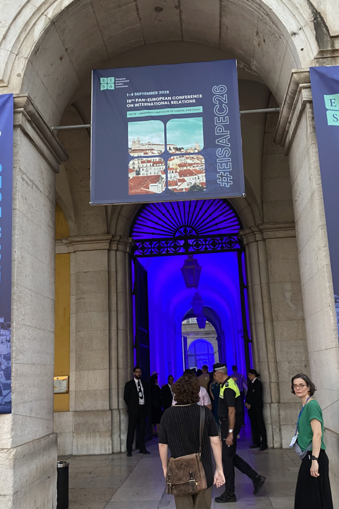 Blick durch einen großen steinernen Torbogen mit blau beleuchtetem Durchgang und mehreren Personen, darunter eine Frau mit grünem Oberteil, davor ein Banner mit der Aufschrift '3rd Pan-European Conference on International Relations 2-4 September 2026 #EISAPEC26'