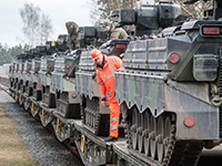 Reihe von Panzern auf Transportfahrzeug, darauf Mann mit orangener Kleidung sowie zwei Bundeswehrsoldaten in Uniform
