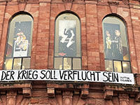 Zu sehen ist das Gebäude des Staatstheaters Mainz mit einem Banner, das die Aufschrift trägt: "Der Krieg soll verflucht sein" 