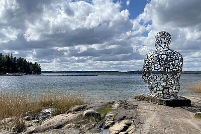 In the foreground is the sculpture “Body of Knowledge” by artist Jaume Plensa, situated on a rock by the shore. The sculpture consists of shiny silver, interconnected letters and depicts a seated human figure with its back turned toward the viewer. A tranquil lake stretches out in the background.