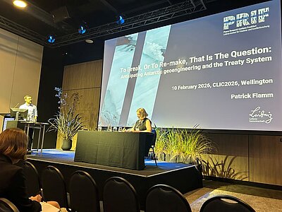 Patrick Flamm auf dem Podium, im Hintergrund eine Präsentation mit dem Titel "To Break, Or To Re-make, That Is the Question: Anticipating Anarctic geoengineering and the Treaty System"
