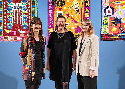 Three women stand in front of a blue wall adorned with colorful tapestries and smile at the camera.