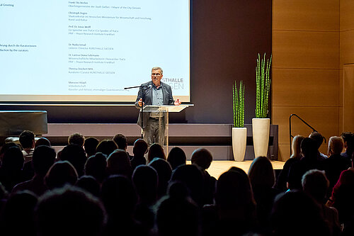 The camera focuses on a man in a suit standing at a lectern on stage, speaking to the audience. A program is projected onto the wall in the background.