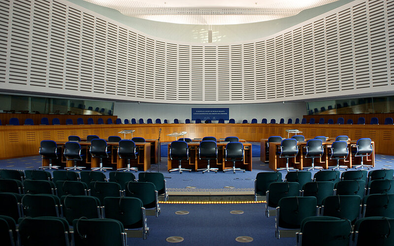 Hall of European Court of Human Rights; blue chairs on the right and left; three tables surrounding the chairs in the center of the picture; in the background, there is a large, oval-shaped wooden platform extending across the room, which serves as a table