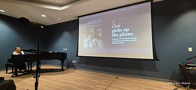 A person is sitting at a grand piano. In the background, Eldad Ben Aharon’s book cover is projected onto a screen.