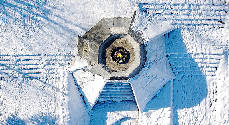 Luftaufnahme des „Eternal Flame Memorial”. Es besteht aus einer Betonplattform mit einer Feuerschale in der Mitte, in der das Feuer brennt. Das Denkmal ist teilweise mit Schnee bedeckt. Auch um das Denkmal herum liegt viel Schnee.