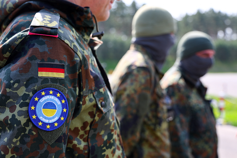 Drei Soldaten. Auf der Uniform des ersten ist eine deutsche Flagge sowie ein Aufnäher mit kombinierter EU Flagge und Ukraine Flagge zu sehen.