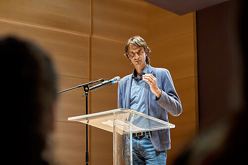 The camera focuses on a man dressed in blue standing at a lectern and speaking to the audience.