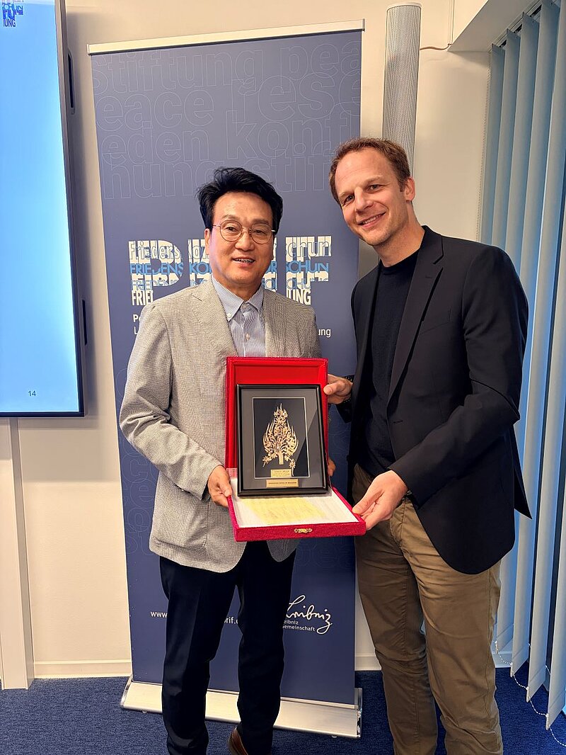 Stefan Kroll steht neben einem Mann, der eine Schachtel mit einem Bild in einem goldenen Rahmen in der Hand hält. Im Hintergrund ist ein Aufsteller mit dem PRIF-Logo zu sehen.