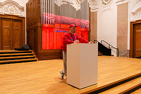 A person dressed in red is standing at a lectern on a stage. A red image is projected onto a screen in the background.