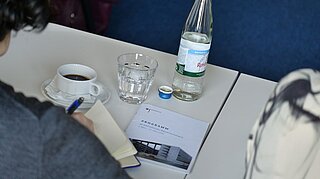 Blick über die Schulter eines Teilnehmenden auf Notizheft, Veranstaltungsprogramm und Kaffeetasse