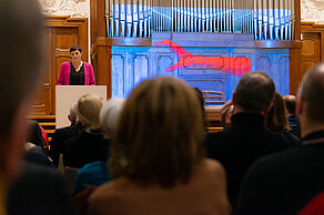 Nicole Deitehoff speaks in front of an audience. A red hand on a blue background is projected onto a screen in the background.