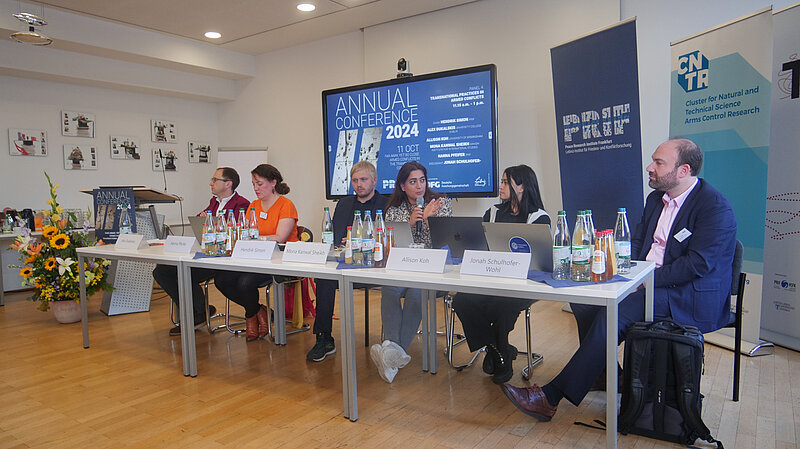 Sechs Personen sitzen auf einem Podium, hinter ihnen ist ein großer Bildschirm auf dem "Annual Conference 2024" in weißer Schrift auf blauem Hintergrund zu erkennen ist.
