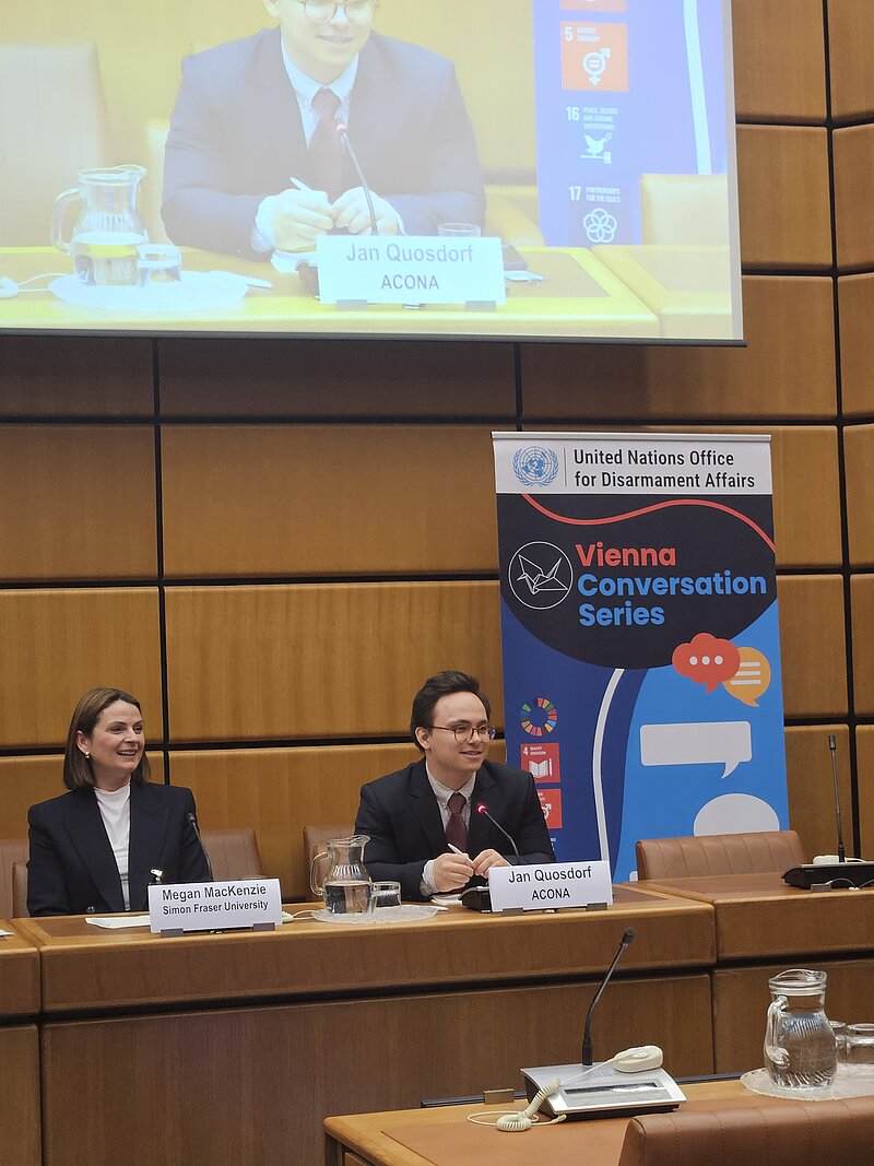 Zwei Menschen sitzen auf einem Podium. Im Hintergrund ist eine Person auf einer Leinwand vergrößert. Daneben steht ein Aufsteller mit dem Logo der Vienna Conversation Series.