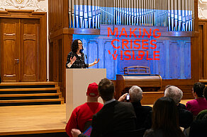 A person stands at a lectern in front of an audience. In the background, the words “Making Crises Visible” are projected onto a screen in red lettering on a blue background.