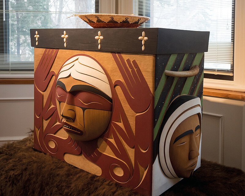 Eine Holzkiste auf der kunstvoll Gesichter abgebildet sind. Die Kiste steht auf einem Fell. Auf der Kiste ist ein Korb.