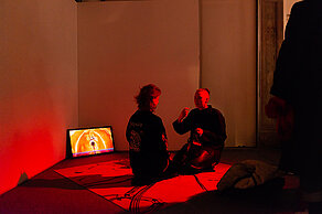 A room is illuminated in red. Two people are sitting on the floor opposite each other. In the background is a screen showing a person running on an orange background.