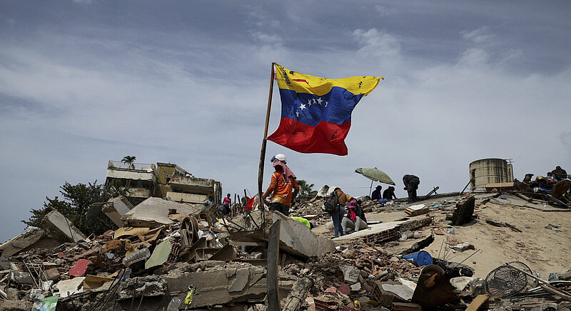 People on rubble with a waving Venezuelan flag under a cloudy sky