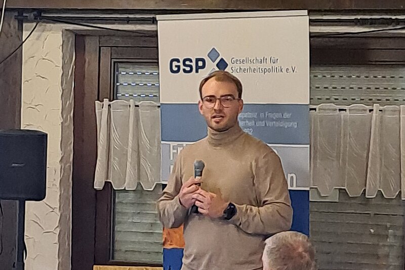 Mikhail Polianskii speaks to an audience with a microphone. A GSP poster can be seen in the background.