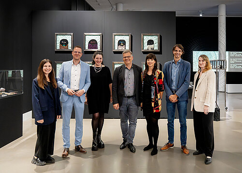 Seven people dressed in formal attire stand in a museum room and pose for the camera.