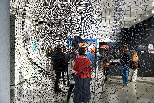 The camera looks into an exhibition space through an art object in the form of a woven spiderweb.
