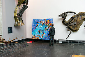 A picture of people wearing life jackets in the water is set up between dinosaur skeletons. A person is standing in front of the picture looking at it.