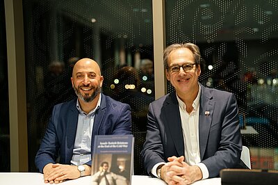 Two men are sitting side by side, smiling at the camera. A book cover can be seen in the foreground.
