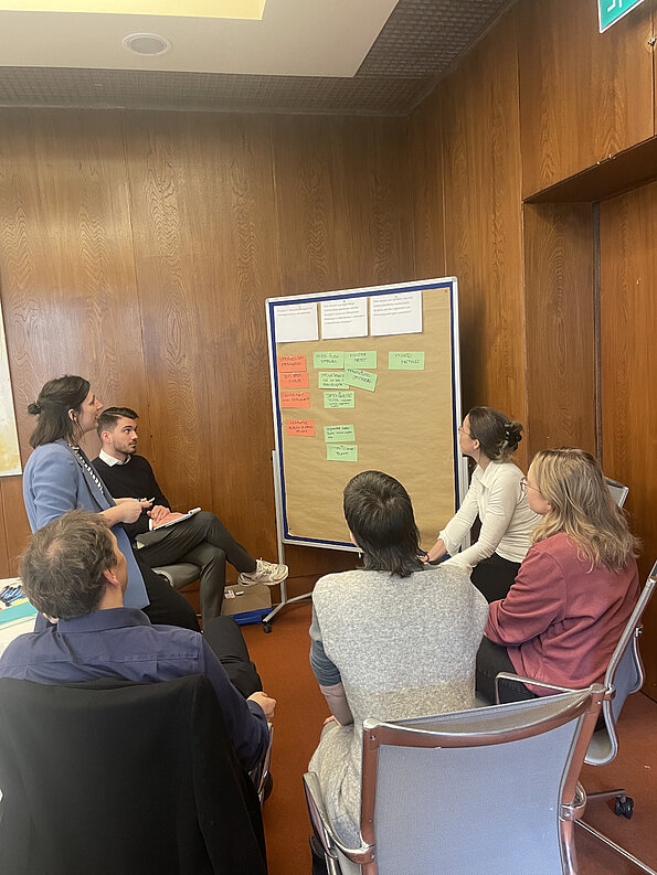 Six people are sitting and standing in front of a board covered with colorful notes.