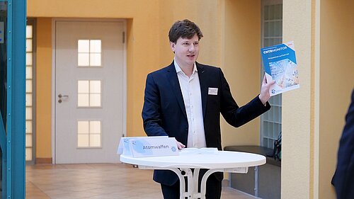 Prof. Malte Göttsche stands at a high table and holds up a blue book entitled CNTR Monitor Perspectives on Dual Use.