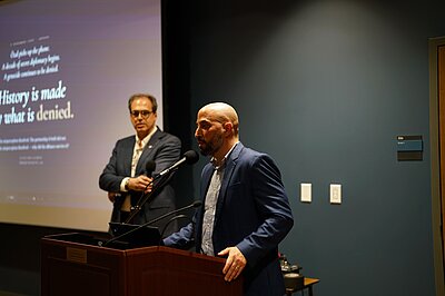 Eldad Ben Aharon is standing at a lectern. Another man can be seen in the background.