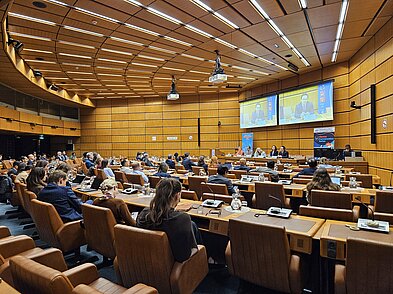 Foto von einem gefüllten Plenarsaal. Auf dem Podium sitzen fünf Menschen. Jan Quosdorf, der auf dem Podium sitzt, ist vergrößert auf zwei Leinwänden im Hintergrund zu sehen.