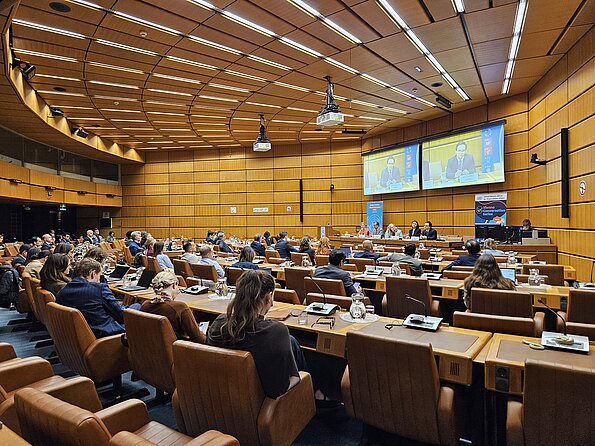 Photo of a packed plenary hall. Five people are seated on the podium. Jan Quosdorf, who is seated on the podium, is shown enlarged on two screens in the background.