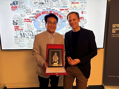 Stefan Kroll steht neben einem Mann, der eine Kiste mit einem goldenen gerahmten Bild in der Hand hält. Im Hintergrund ist auf einem Screen eine Illustration zum Global House of Young Voices zu erkennen.