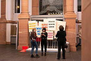 Three people stand in front of signs on which words are depicted in different colors: “Framing, Blaming, Gaming”, ‘Relation, Rotation, Migration’, ”Proceed, Succeed, Repeat”