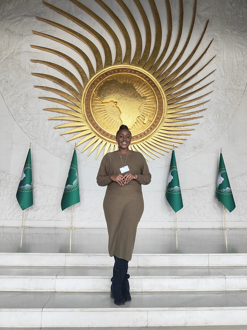 Hilda Koyier in der AU Meeting Hall. Im Hintergrund eine goldene Sonne und grüne Flaggen.