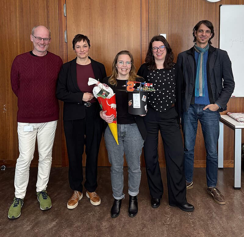 Fünf Personen stehen in einer Reihe und lächeln in die Kamera, darunter Nicole Deitelhoff, Diane Schumann und Jonas Wolff. Dina Schumann steht in der Mitte und hält einen verzierten Doktorhut und eine Schultüte in der Hand.