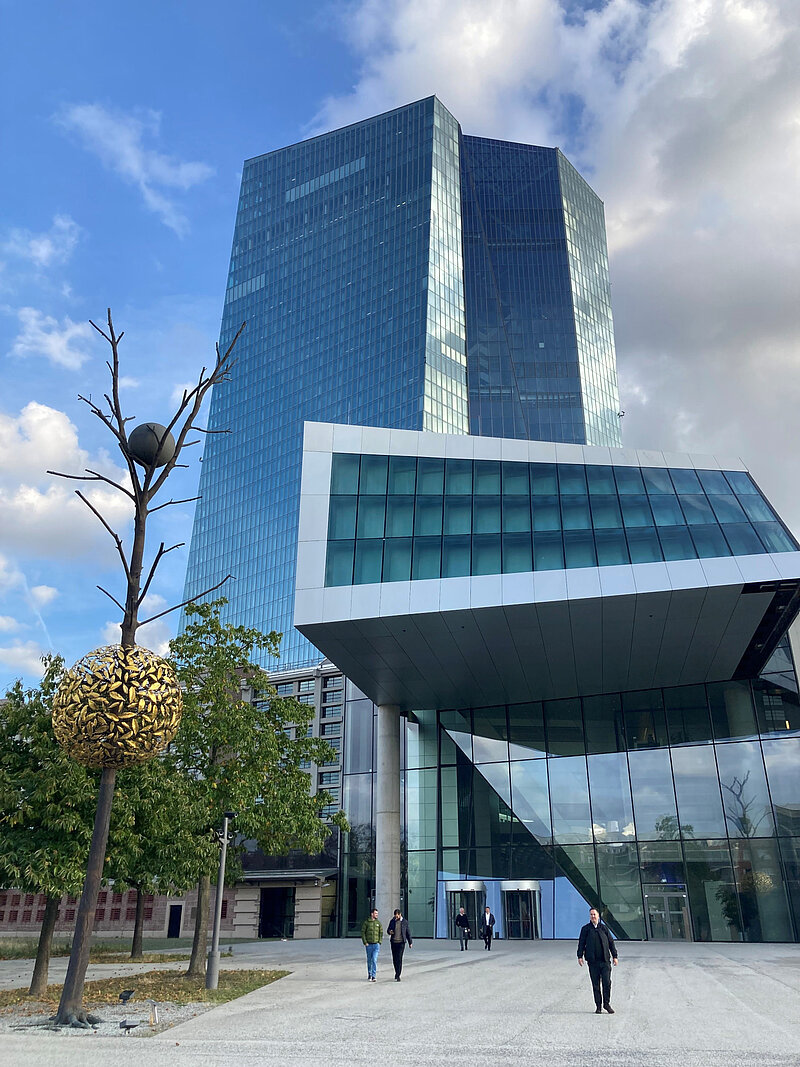 Der Nordeingang der EZB in Frankfurt mit dem markanten gläsernen Eingangsgebäude und dem hohen, spiegelnden Hauptturm im Hintergrund, davor eine Skulptur mit goldener Kugel an einem kahlen Baumstamm.