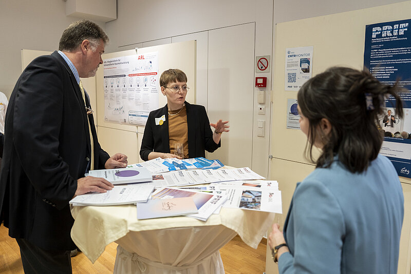 Drei Personen stehen an einem Stehtisch, auf dem PRIF-Publikationen ausliegen, und sprechen miteinander. Im Hintergrund hängt ein Poster mit dem PRIF-Logo an der Wand.