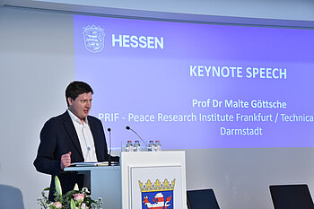 Malte Göttsche is standing at a lectern bearing the coat of arms of Hesse. A slide from the presentation can be seen in the background.