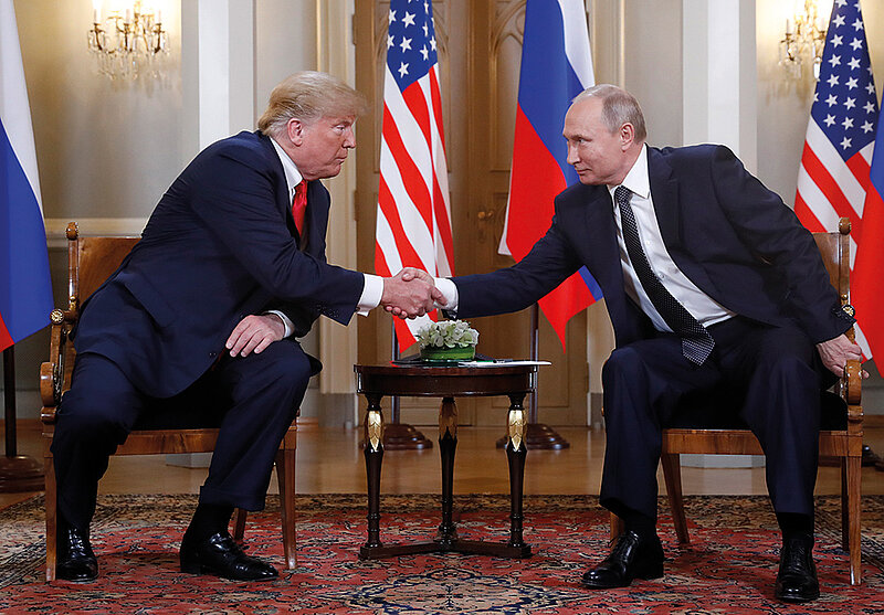 zwei Männer schütteln einander sitzend die Hand. Im Hintergrund die US Flagge und die russische Flagge.