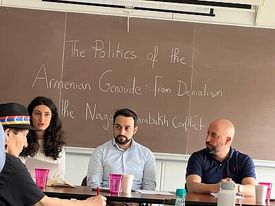 Four people are sitting at tables. In the background, a sign reads “The Politics of the Armenian Genocide: From Denialism to the Nagorno-Karabakh Conflict”