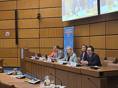 Fünf Menschen sitzen auf einem Podium. Im Hintergrund sind Leinwände und ein Aufsteller von United Nations Office for International Affairs zu sehen.