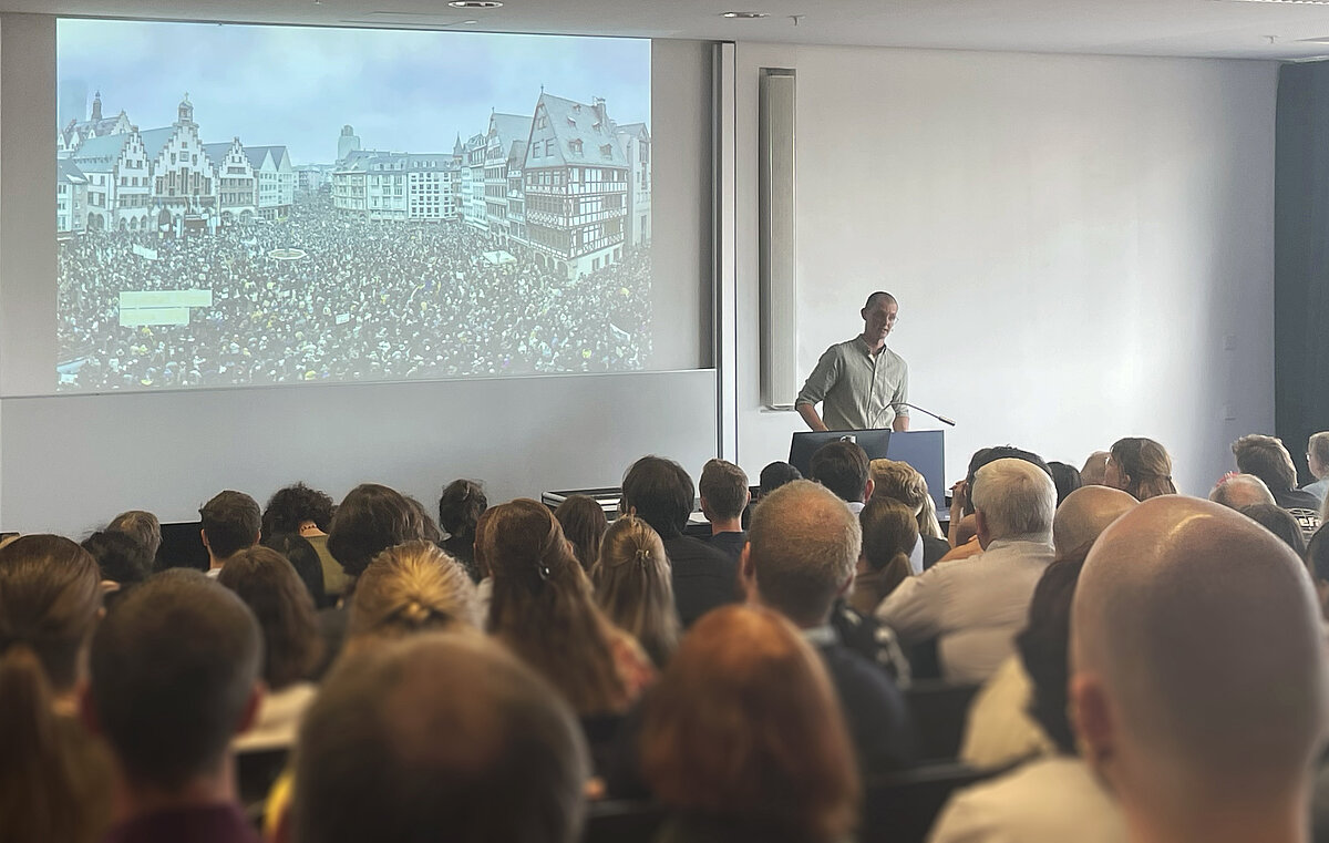 Gefüllter Raum, ein Mann vorne am Rednerpult, an die Wand projiziert ein Foto vom mit Menschen gefüllten Frankfurter Römer