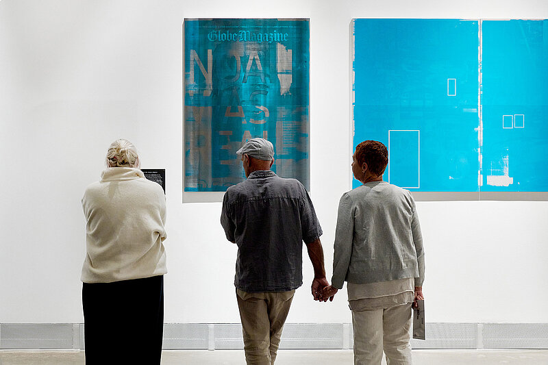 Drei Personen stehen nebeneinander und schauen auf zwei blaue Bilder in einer Ausstellung. Zwei der Personen halten sich an den Händen.