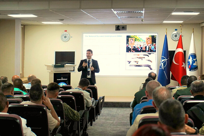 Jonatan Kurzwelly steht vor einer Präsentationsfolie und spricht vor Publikum. Rechts von ihm sind die Flaggen der NATO, der Türkei und des COE-DAT zu erkennen.
