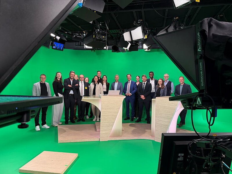 Eine Gruppe von 17 Personen steht vor einem Greenscreen in einem Fernsehstudio