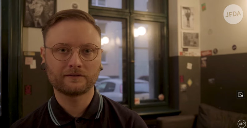 Ausschnitt aus einer Doku. Constantin Winkler sitzt in einem Raum und schaut in die Kamera. 