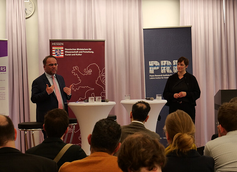 Timon Gremmels und Nicole Deitelhoff stehen vor einem Publikum. Im Hintergrund stehen Aufsteller mit dem Logo von PRIF und dem Hessischen Ministerium für Wissenschaft und Forschung, Kunst und Kultur