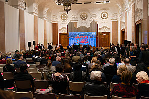 Many people are sitting in the audience, some people are still standing. The words “Making Crises Visible” are projected onto a screen on the stage in red lettering on a blue background.